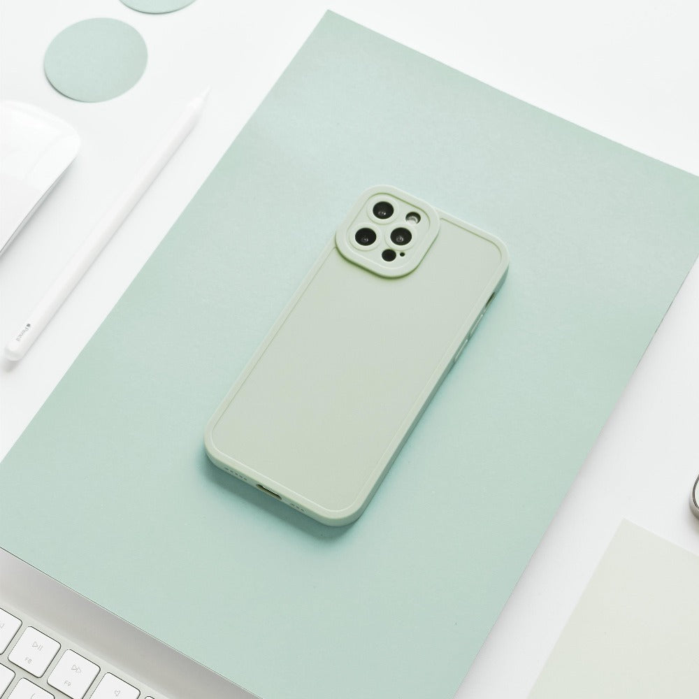 A sage green smartphone with a matching NAKD Phone Case - Sage by Coconut Lane lies on a mint green surface, surrounded by minimalist office supplies including a white keyboard, a white pen, and mint green circular stickers, creating a monochromatic aesthetic.