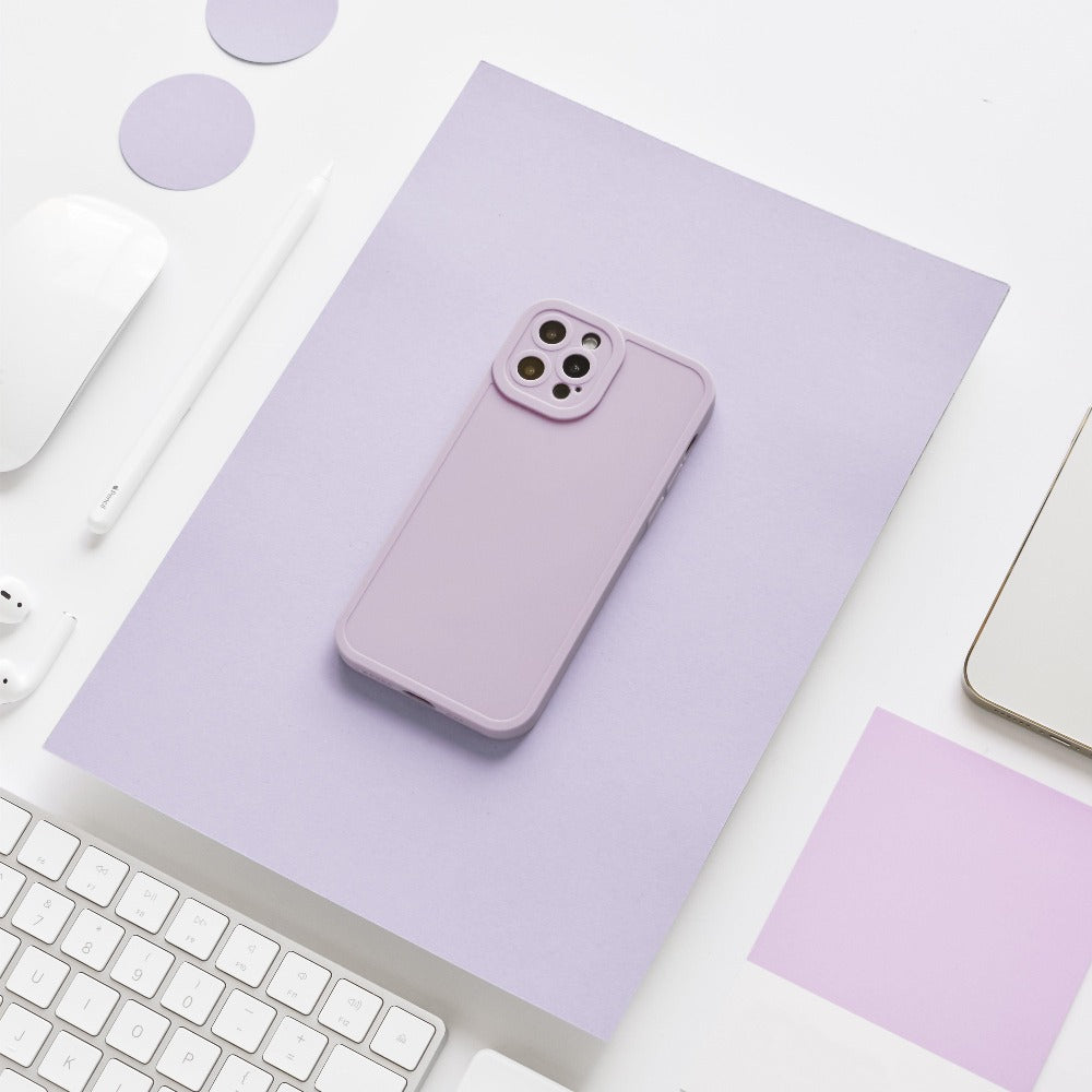 A lilac-colored smartphone case from the Coconut Lane NAKD collection is placed on a matching lilac sheet of paper. Surrounding it are various office supplies, including a white keyboard, a white pencil, a white wireless earbud, and a partially visible laptop. The setting is minimalistic and clean.