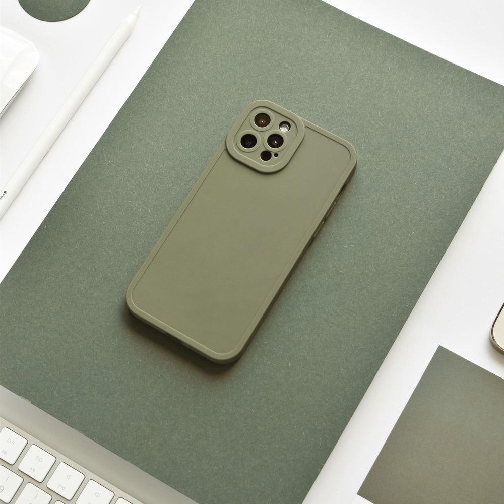 A NAKD Phone Case - Khaki by Coconut Lane is placed on a green rectangular mat on a white desk. Nearby, there is a white keyboard, a white pencil, and part of a white phone edge visible. The full camera protection and overall setting suggest a minimalist and organized workspace.