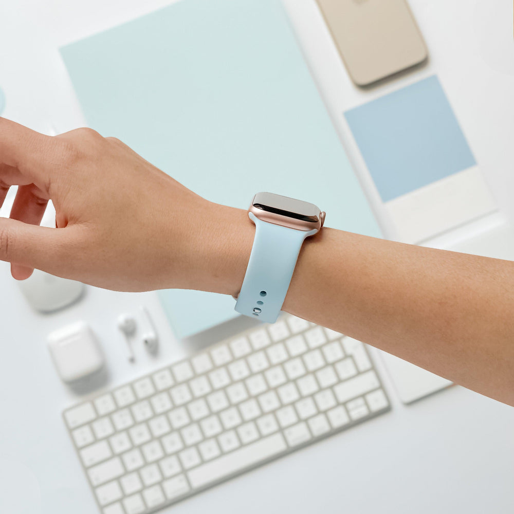A person's arm is shown wearing the Coconut Lane NAKD Apple Watch Strap in Sky Blue. The background includes a white keyboard, wireless earbuds, and various sleek tech accessories in shades of white and spring-inspired pastels. The setting appears to be a clean and modern workspace.