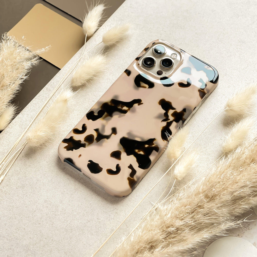 A smartphone with an Ivory Tort Phone Case from Coconut Lane is placed on a beige surface surrounded by soft, fluffy pampas grass. The phone's triple camera setup is visible, and there is a hint of a laptop or tablet in the background.
