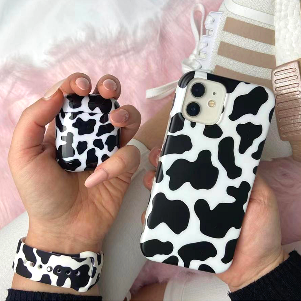 Close-up of a person holding a black and white cow-print AirPods case along with a matching iPhone case. Wearing Coconut Lane's Cow Apple Watch Strap on their left wrist, they complete the look with white sneakers featuring beige accents. The background showcases a soft pink surface.