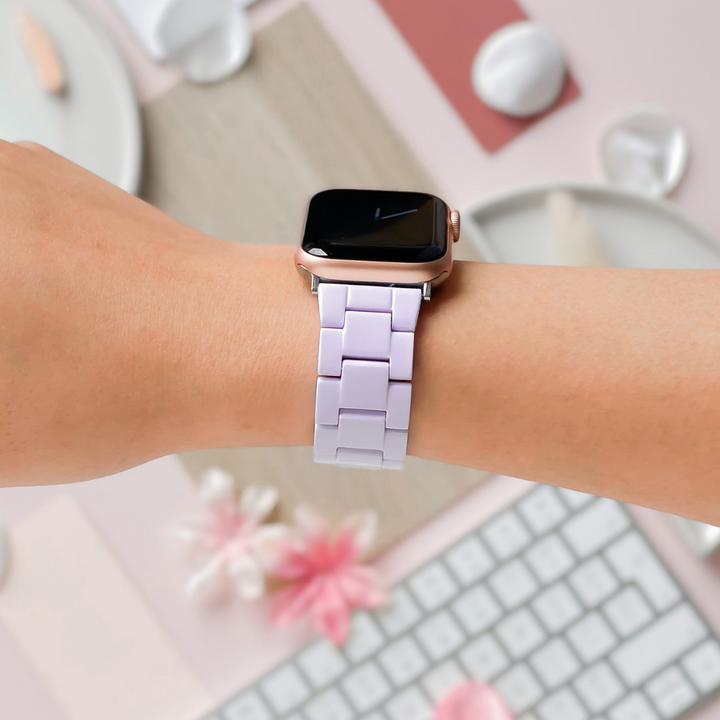 A close-up of a person wearing a smartwatch featuring the Luxe White Apple Watch Strap by Coconut Lane on their left wrist. The background includes a keyboard, a pink flower, and some decorative items in pastel colors, crafting a soft, aesthetically pleasing setting.