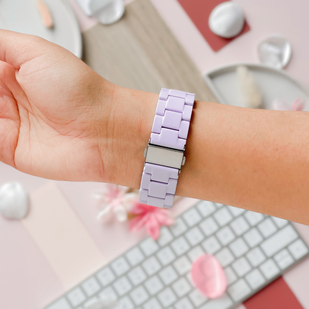 A person shows off their wrist adorned with the Luxe White Apple Watch Strap by Coconut Lane, featuring an adjustable, lavender-colored strap and a metal clasp. The background includes a keyboard, decorative stones, and pastel-colored items, creating a soft, aesthetically pleasing composition.