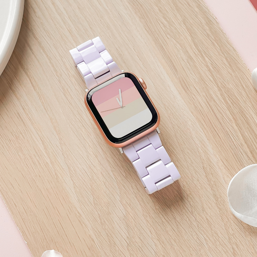 A smartwatch featuring the Luxe White Apple Watch Strap from Coconut Lane, with a rose gold casing and a pastel pink and lavender striped adjustable strap, lies on a light wood surface. The screen displays a minimalistic clock face with pastel shades that complement the band. Delicate white accessories are partially visible in the corners.
