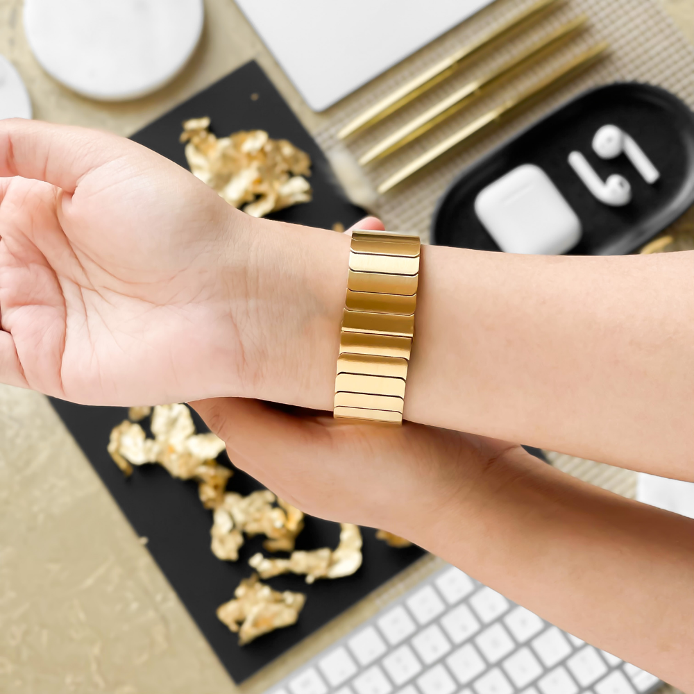 A person is trying on the Coconut Lane Slinky Apple Watch Strap in Gold, adjusting it on their wrist. The background features a desk with scattered golden foil pieces, a keyboard, pens, and AirPods in their case. The overall color scheme is gold and white.