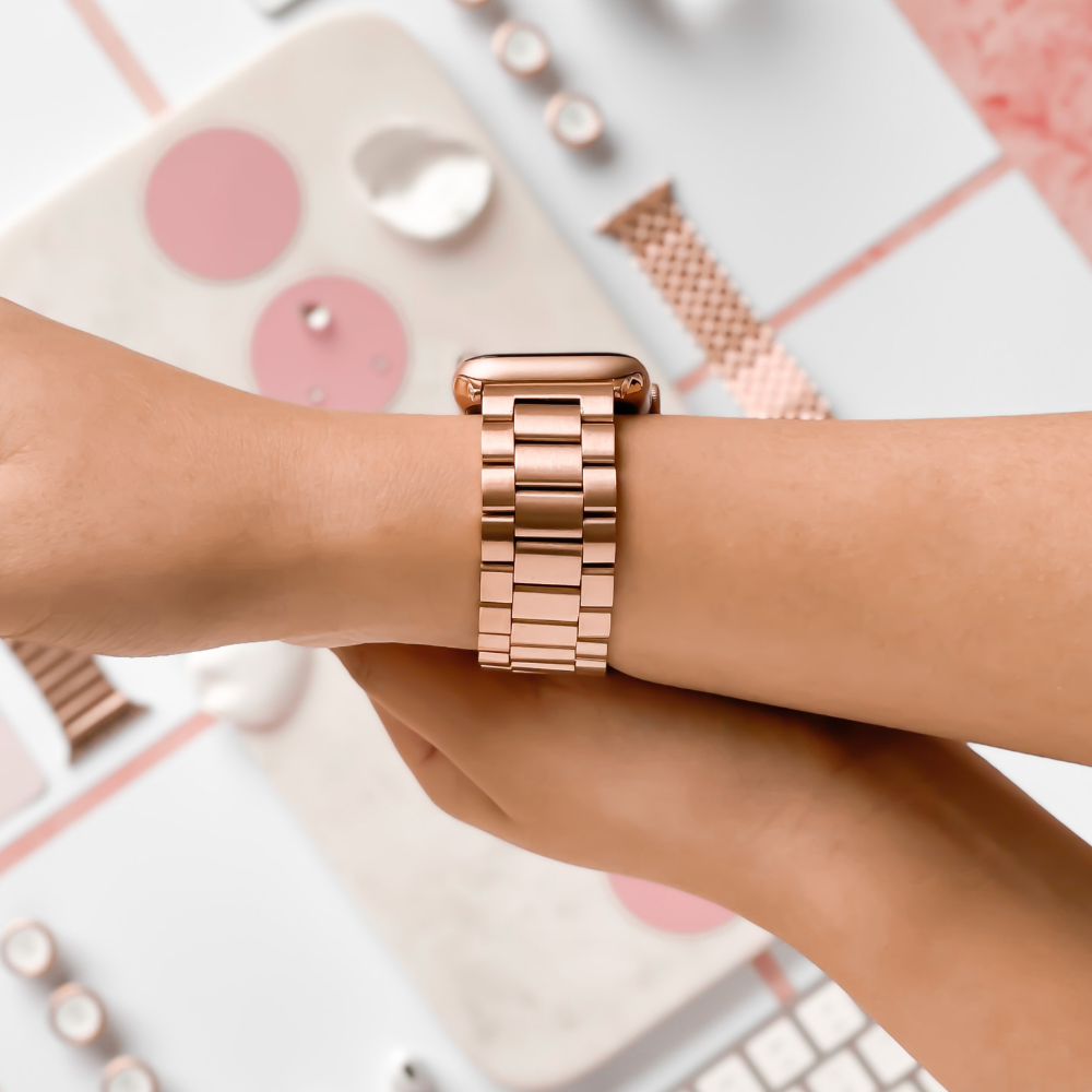 Close-up of a person's wrist wearing a Coconut Lane Stainless Steel Apple Watch Strap in Rose Gold. The background features various pink and white objects, including other watch bands and circular decorations, all arranged on a flat surface. Perfect for all Apple Watch models, this strap is both sleek and stylish.